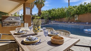 Un espace extérieur aménagé pour les repas, avec un barbecue, une piscine et des palmiers.