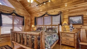 A log cabin bedroom with a wooden bed, two windows with blinds, a nightstand, a chair, and a painting on the wall. - Bear-riffic View | Hot Tub, Games & Scenic Decks (Sevierville)