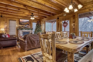 A cozy log cabin interior with a fireplace, leather sofas, a dining table with placemats, and a Christmas tree.
