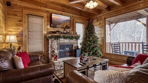 A cozy log cabin interior with a fireplace, a Christmas tree, leather furniture, and a television mounted on the wall. - Bear-riffic View | Hot Tub, Games & Scenic Decks (Sevierville)