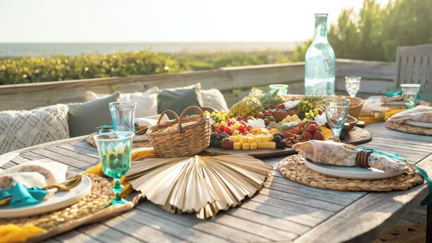 A wooden table set with a variety of foods, including fruits, cheeses, and bread, with a view of the ocean in the background. - Jonah's Whale: Large Oceanfront Home, Ocean and Waterway Views (Caswell Beach)