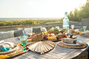 Uma mesa de madeira posta com diversos alimentos, incluindo frutas, queijos e pão, com vista para o oceano ao fundo.