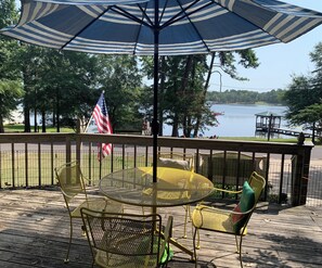 A deck with a round table and chairs, an American flag, and a view of a lake. - Lakehouse, Sleeps 6, Private Dock, Infinity Beach Pit, 2 Kayaks! (Hawkins)
