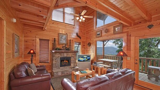 A cozy cabin interior with a fireplace, leather sofas, wooden furniture, and large windows offering views of the outdoors. - Time Well Wasted | Hot Tub, Game, Loft, Resort Pool, Views & Family-Friendly (Sevierville)