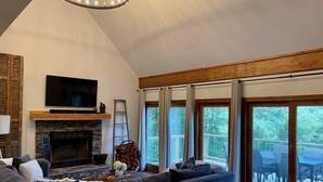 Een ruime woonkamer met een hoekbank, een open haard, een aan de muur bevestigde televisie en grote ramen met uitzicht op het omliggende landschap.