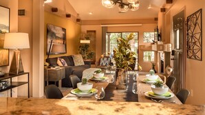 A modern dining area with a long table set for six, surrounded by chairs. There is a sofa and a small side table with a lamp in the background. - Jewel House West Sedona with Private Hot Tub! (Sedona)