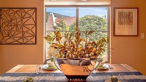 A dining area with a table set for two, a vase with dried plants, and a window with a view of a red-roofed building and greenery. - Jewel House West Sedona with Private Hot Tub! (Sedona)