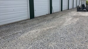 Uma longa fileira de portões de garagem brancos fechados, com detalhes verdes, um caminho de cascalho e um veículo estacionado com uma reboque.