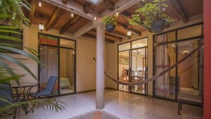 A spacious indoor area with a tiled floor, a central column, and hanging plants. There are glass doors leading to another room with a dining table and chairs.