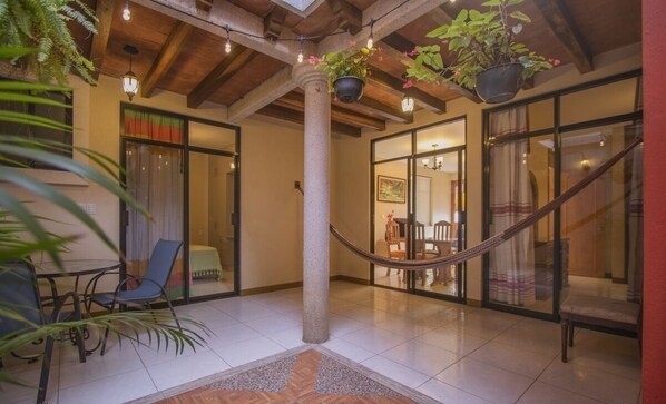 Amplio espacio interior con suelo de baldosas, una columna central y plantas colgantes. Hay puertas de cristal que dan a otra habitación con una mesa y sillas para comer.
