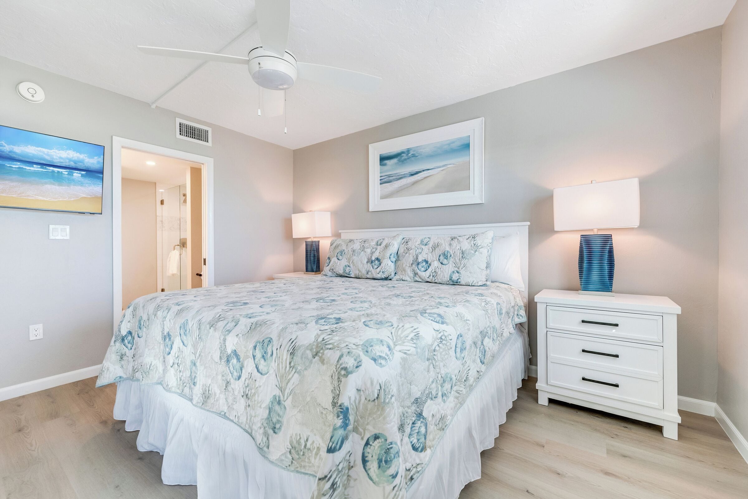 Een slaapkamer met een bed, bedlampjes, een nachtkastje, een plafondventilator en een schilderij met een strandthema aan de muur.