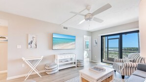 Una sala de estar moderna con un gran ventanal, un ventilador de techo y vista a la playa.