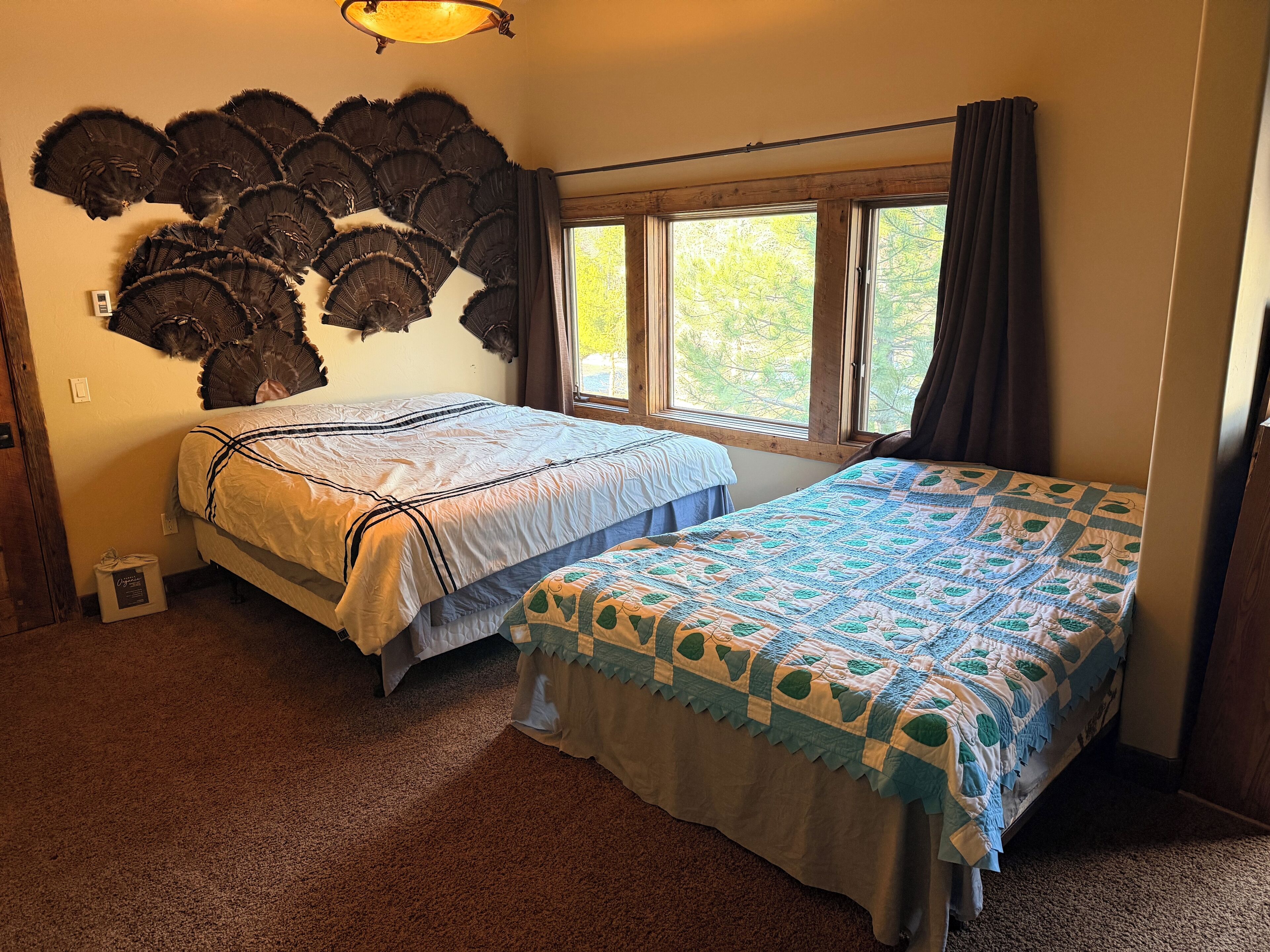 A room with two beds, a wall-mounted fan, a window with curtains, and a carpeted floor.