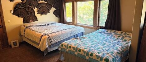 A room with two beds, a wall-mounted fan, a window with curtains, and a carpeted floor.