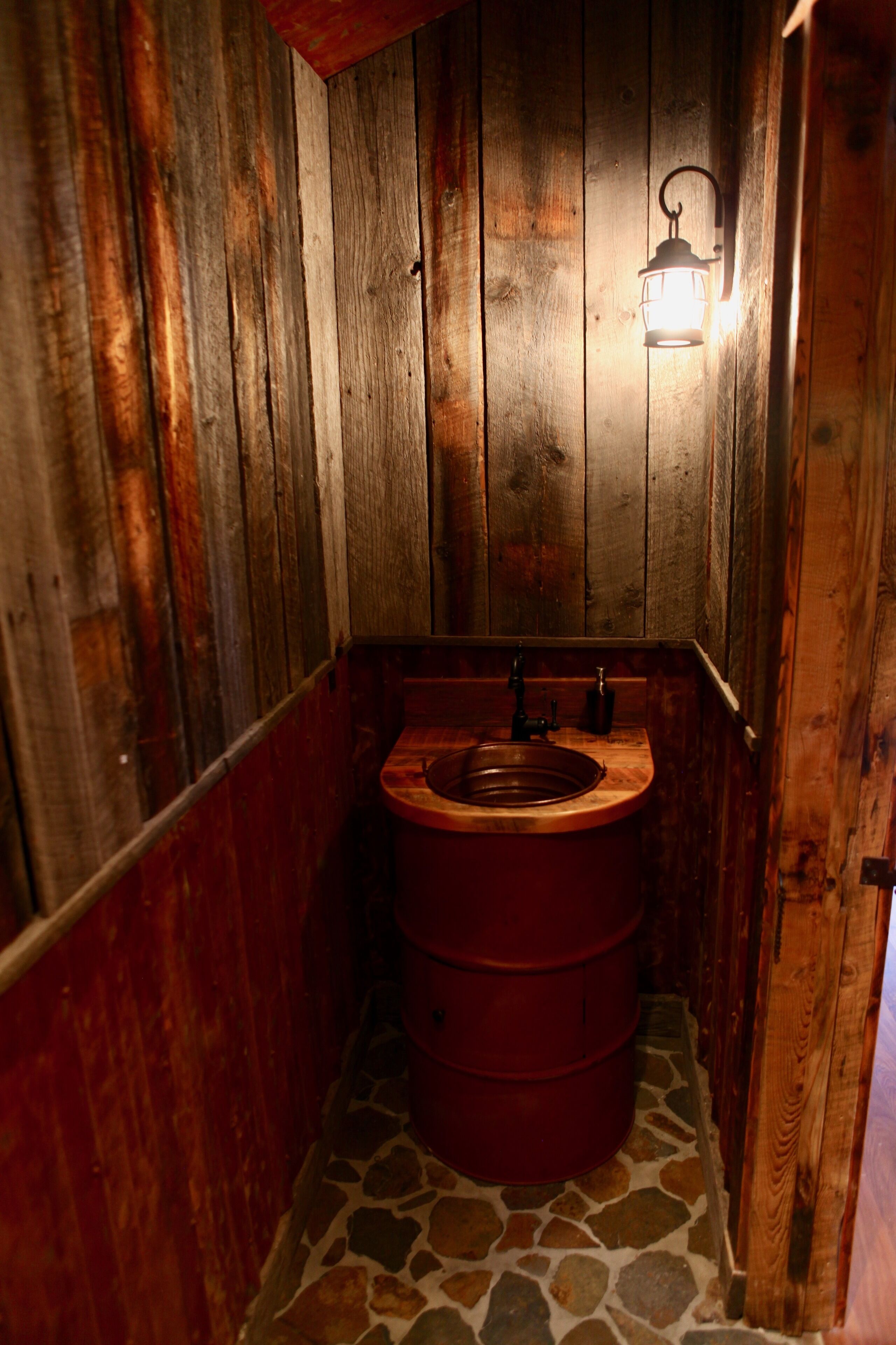 Une salle de bain rustique en bois, avec un sol carrelé en pierre, des toilettes en bois et un luminaire fixé au mur.