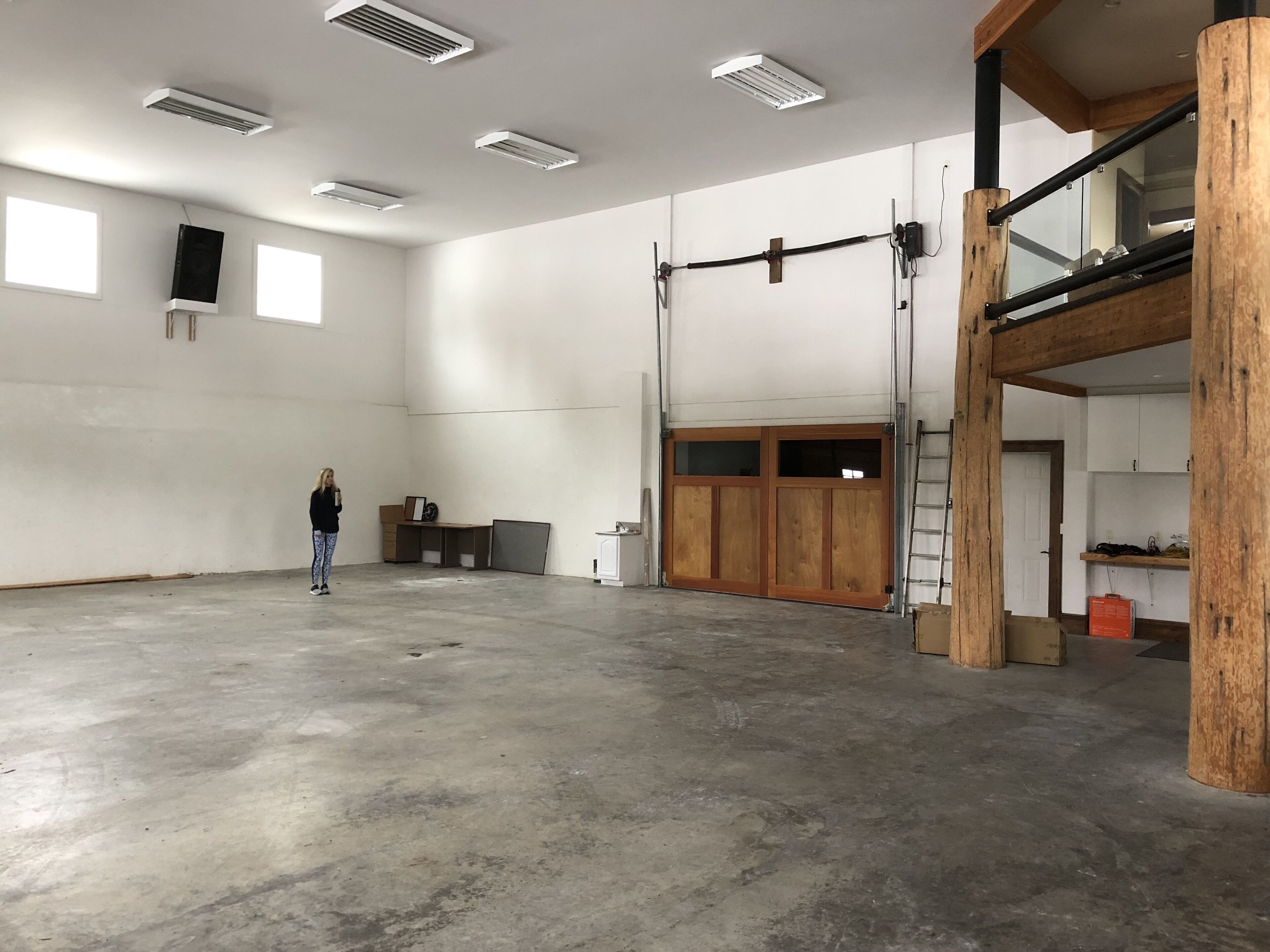 Um espaço amplo e interno, com piso de concreto, vigas de madeira e uma grande porta deslizante.