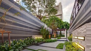 Un edificio moderno con fachada elegante y a rayas, un jardín bien cuidado con diversas plantas y un camino que lleva a una zona cubierta.