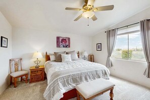 Une chambre à coucher comprenant un lit, une chaise, une table de chevet, une lampe, un ventilateur de plafond, une fenêtre avec des rideaux et un tableau accroché au mur.