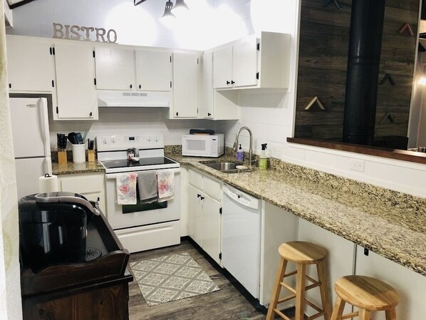 Una cocina con alacenas blancas, estufa, microondas y cafetera. Hay dos taburetes y una alfombra en el piso.