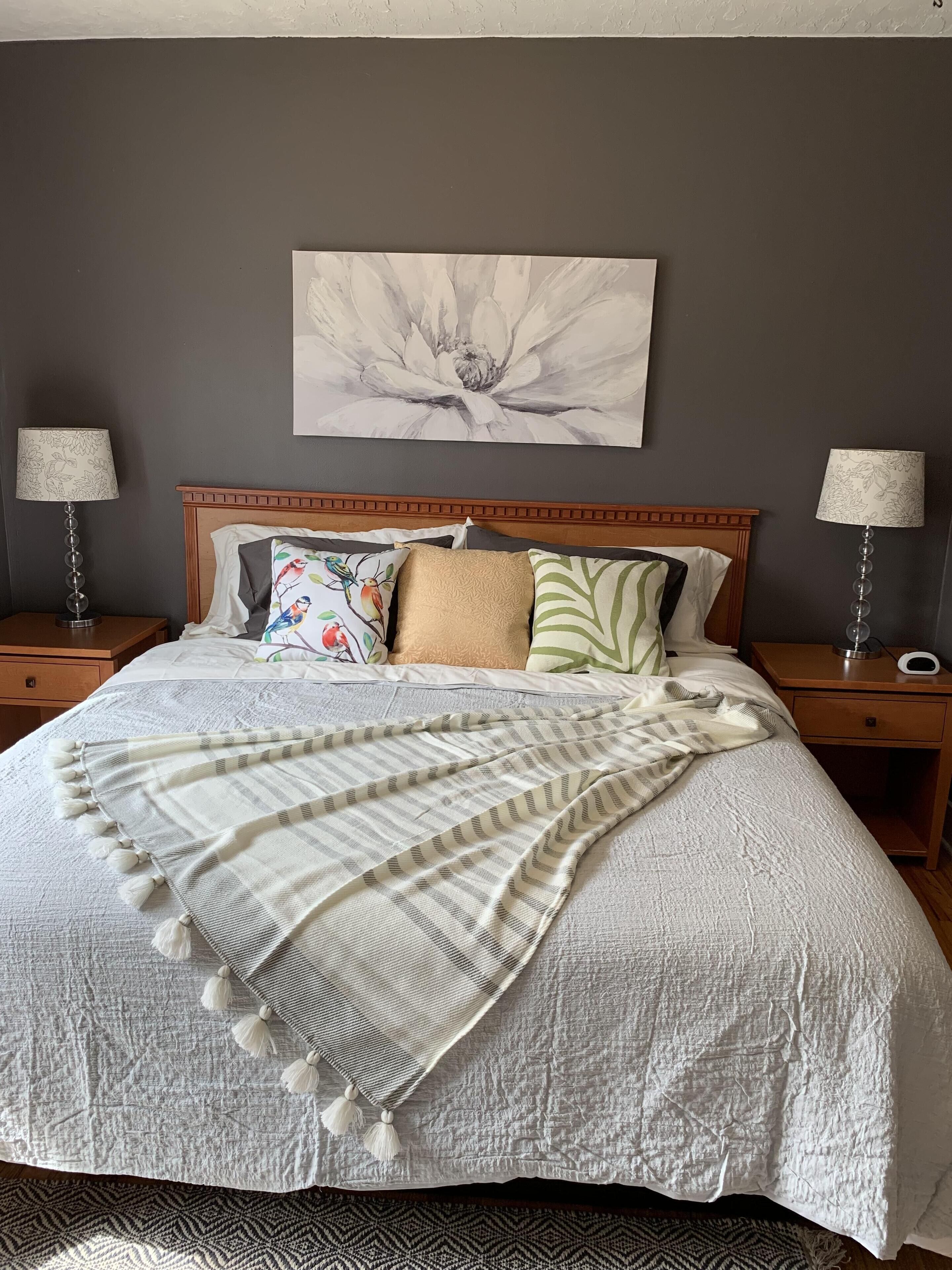Un lit bien fait, agrémenté d’un coussin à motifs floraux, d’un plaid rayé et de deux lampes de chevet posées sur des tables de nuit en bois.