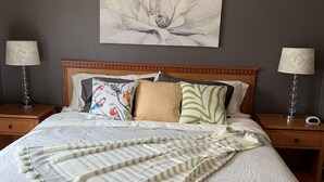 Een net opgemaakt bed met een kussentje met bloemenprint, een gestreept plaid en twee bedlampjes op houten nachtkastjes.