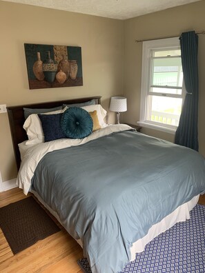 Une chambre à coucher avec un grand lit, une table de chevet, une lampe, une fenêtre avec des rideaux et un tableau au mur.