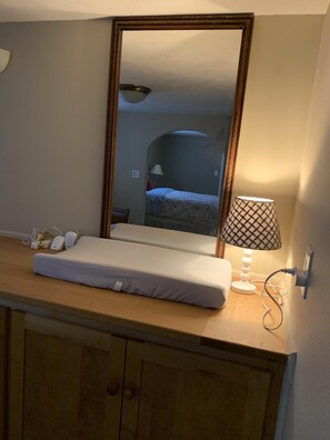 Une chambre d’hôtel avec une coiffeuse en bois, un miroir, une lampe de chevet, un matelas blanc et une table de chevet avec une lampe et des câbles de recharge.