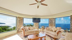 A spacious living room with two sofas, a coffee table, and a television. The room has large windows offering a view of the ocean. - 🌴Joy Estate by IRE Vacations - Expansive Property with Amazing Views ☀️ (Cay Hill)