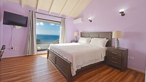 A bedroom with a wooden bed, a nightstand with a lamp, a TV mounted on the wall, and a view of the sea through a large window. - 🌴Joy Estate by IRE Vacations - Expansive Property with Amazing Views ☀️ (Cay Hill)