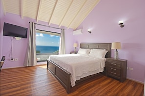 Une chambre avec un lit en bois, une table de chevet avec une lampe, un téléviseur fixé au mur et une vue sur la mer par une grande fenêtre.