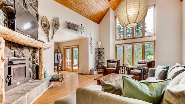 A spacious living room with a stone fireplace, wooden beams, and large windows offering views of the outdoors.