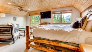 A bedroom with a wooden ceiling, a bed with white bedding, a mounted TV, and large windows with a view of trees and mountains.