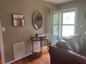 A room with a wooden floor, a brown couch, a small wooden table with a decorative mirror, a framed picture on the wall, and a door leading outside.