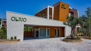 Een modern gebouw met de naam 'OLIVO' op de gevel, met een combinatie van glas en houten elementen, en een bestraat erf met beplanting.