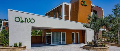A modern building with the name "OLIVO" on the facade, featuring a mix of glass and wooden elements, and a paved courtyard with landscaping.