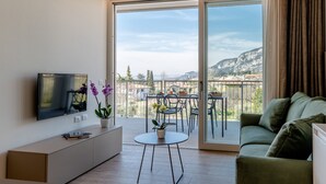 Een moderne woonkamer met een flatscreen televisie, een bank, een klein rond tafeltje met een potplant en een balkon met een tafel en stoelen.