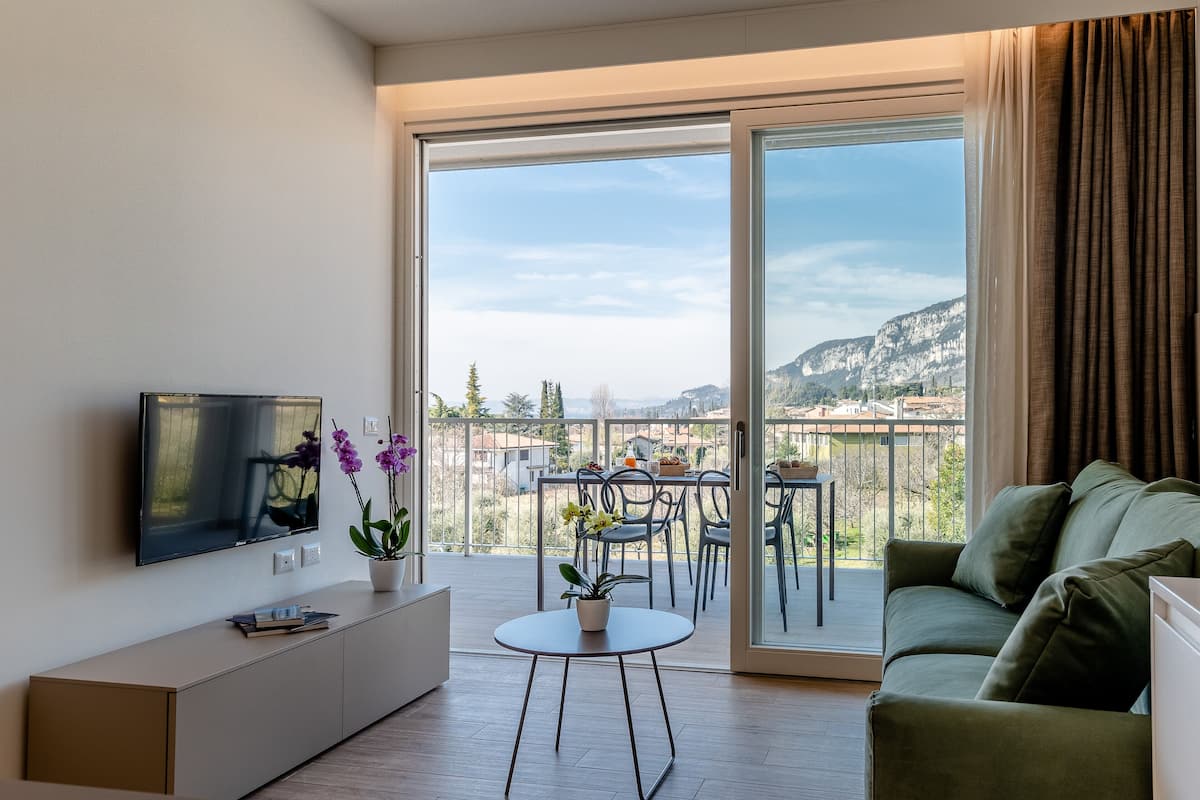 Ein modernes Wohnzimmer mit Flachbildfernseher, einer Couch, einem kleinen runden Tisch mit Topfpflanze und einem Balkon mit Tisch und Stühlen.