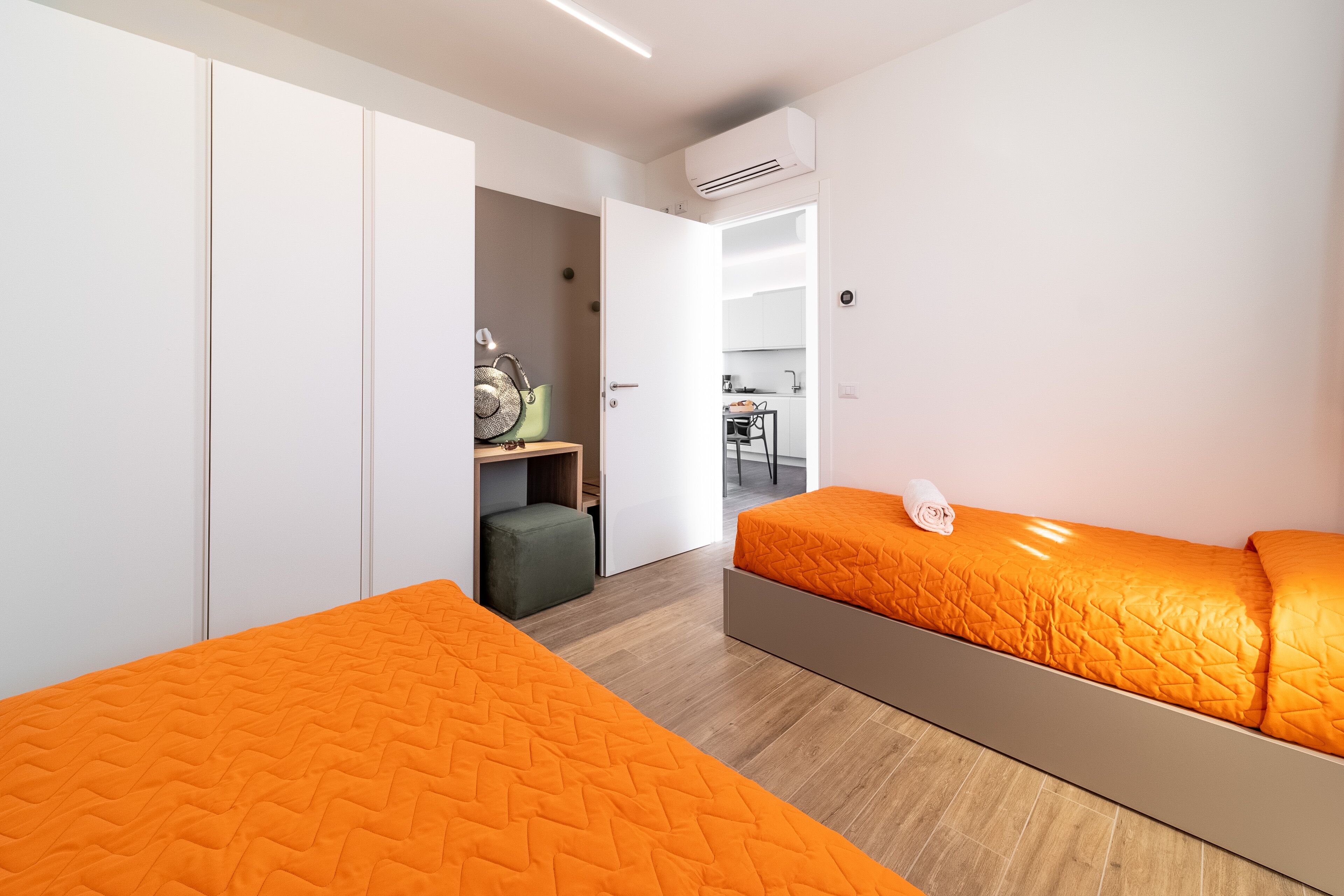 A modern bedroom with two beds, a wardrobe, a kitchenette, and an open door leading to a dining area.