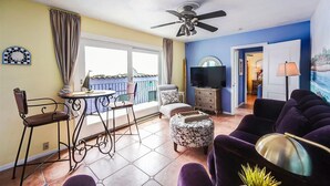 Ein Wohnzimmer mit Balkon, Deckenventilator, Fernseher und Essbereich mit Tisch und Stühlen.
