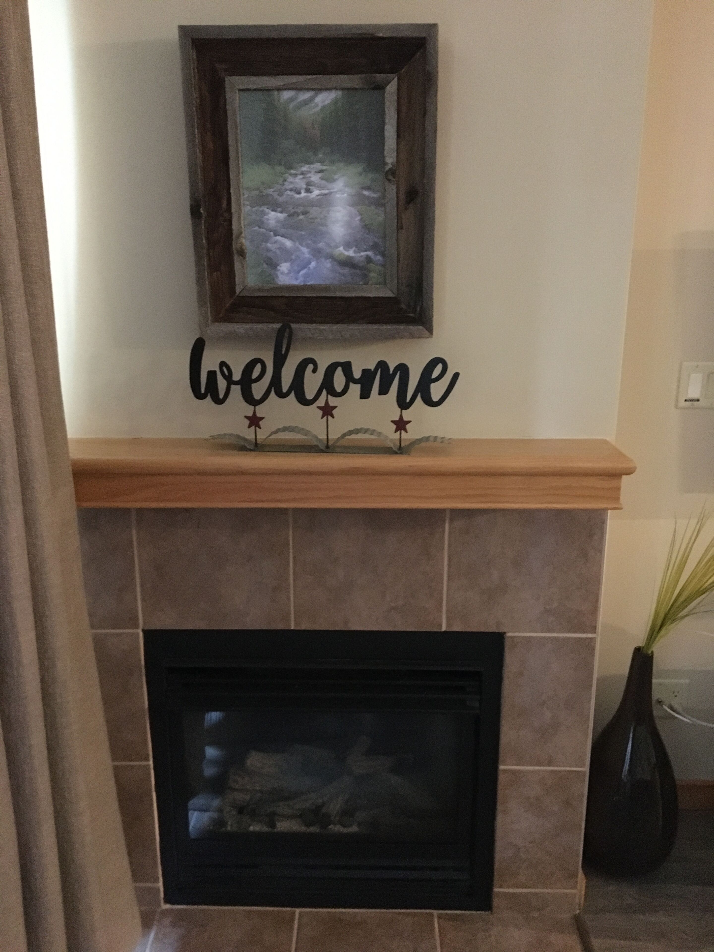 A fireplace with a welcome sign above it, a framed picture on the wall, and a vase with plants to the right.
