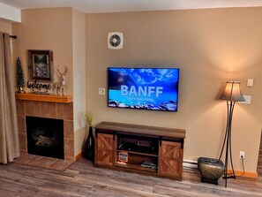 Un salon avec un téléviseur affichant l’image du « PARC NATIONAL DE BANFF », une cheminée, un meuble de rangement en bois et une lampe.
