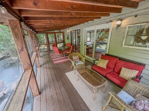 A covered porch with wooden flooring, red and wicker furniture, and large windows offering a view of the outdoors. - RIVER FRONT HOUSE INSIDE THE COOSAWATTEE RESORT (Ellijay)