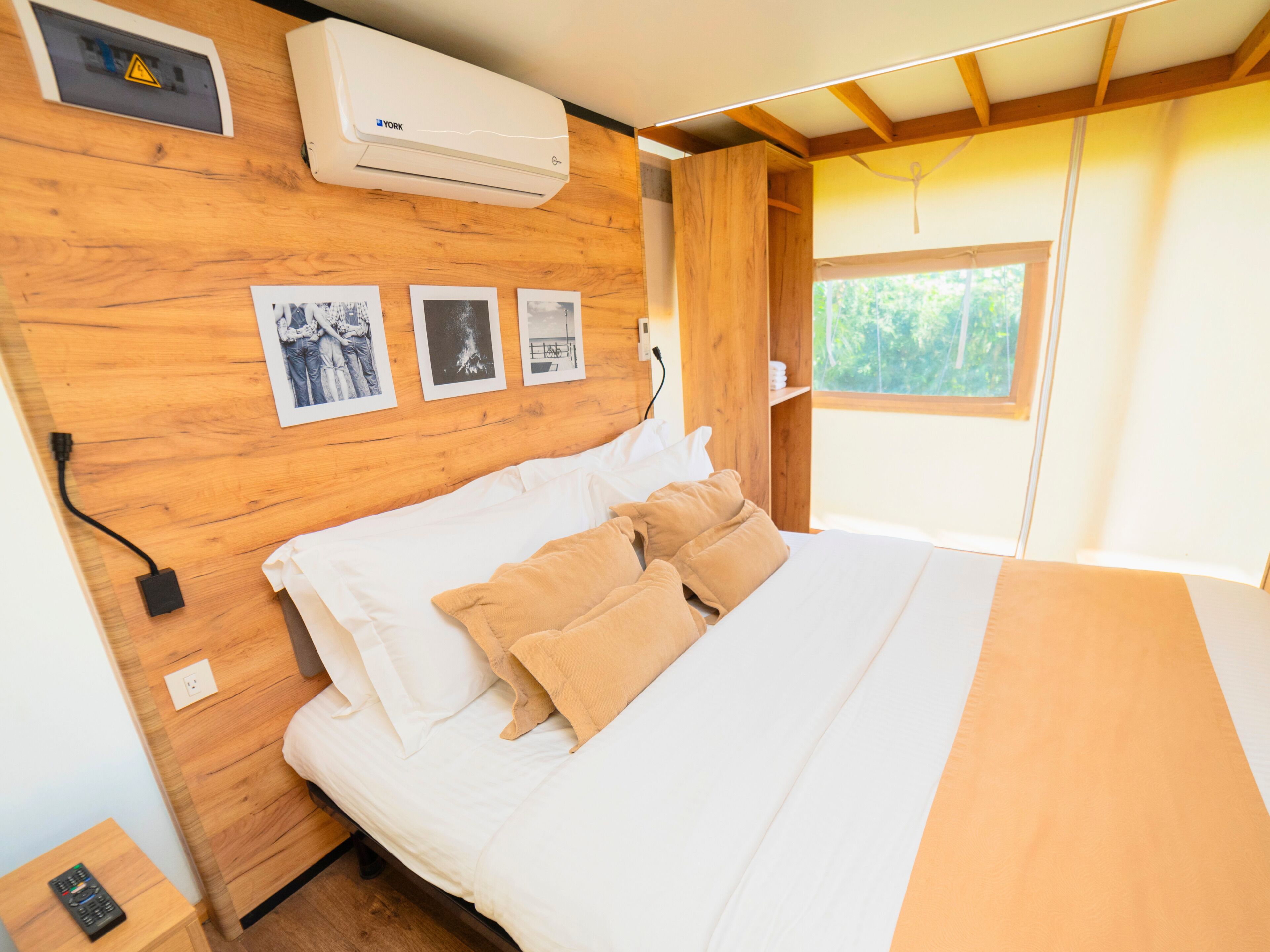 A bedroom with a wooden headboard, a bed with white and beige bedding, a wall-mounted air conditioner, and a window with a view of greenery.