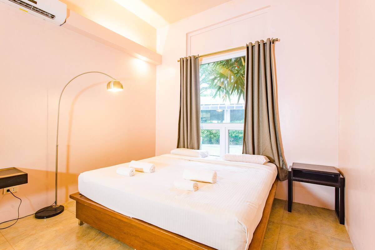 A hotel room with a bed, a bedside table, a floor lamp, and a window with curtains.
