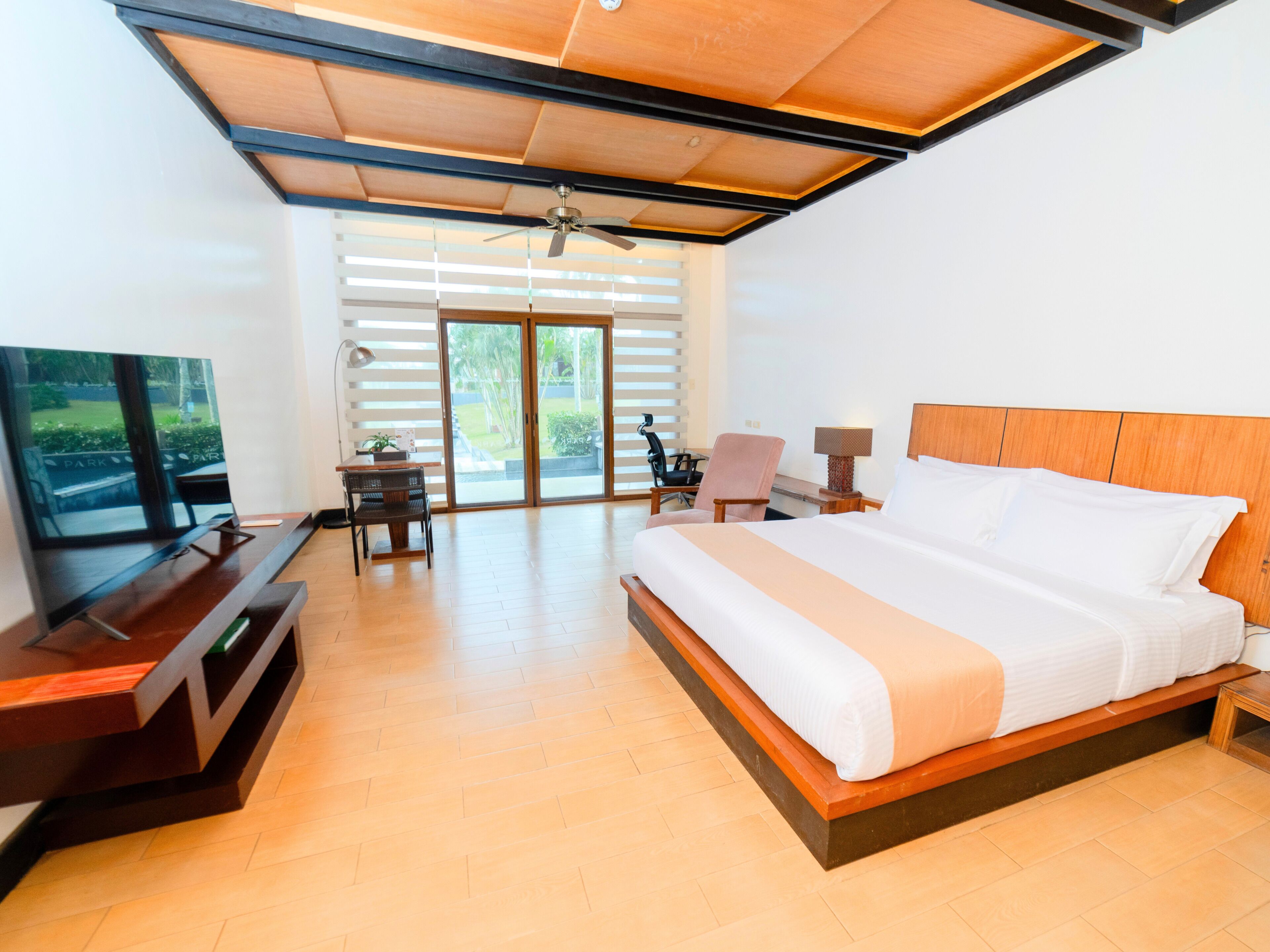 A modern hotel room with a large bed, a desk, a chair, a television, and a view of a park through the sliding doors.