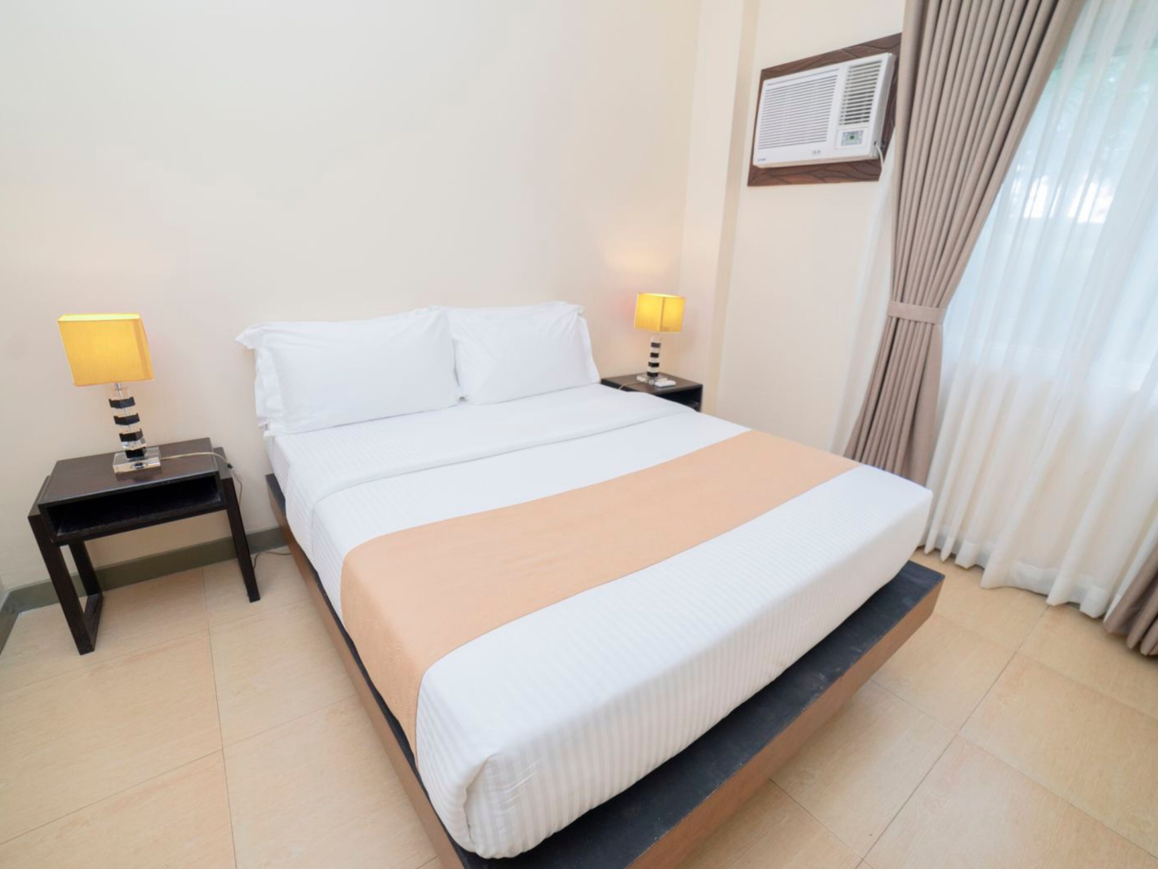 Hotelkamer met een bed, twee nachtkastjes met lampen, een raam met gordijnen en een airconditioner aan de muur.