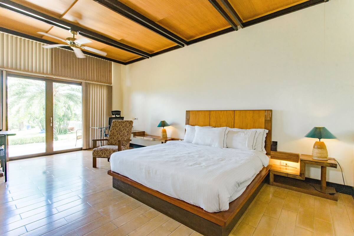 A spacious bedroom with a large bed, a ceiling fan, and a view of palm trees through the sliding glass doors.