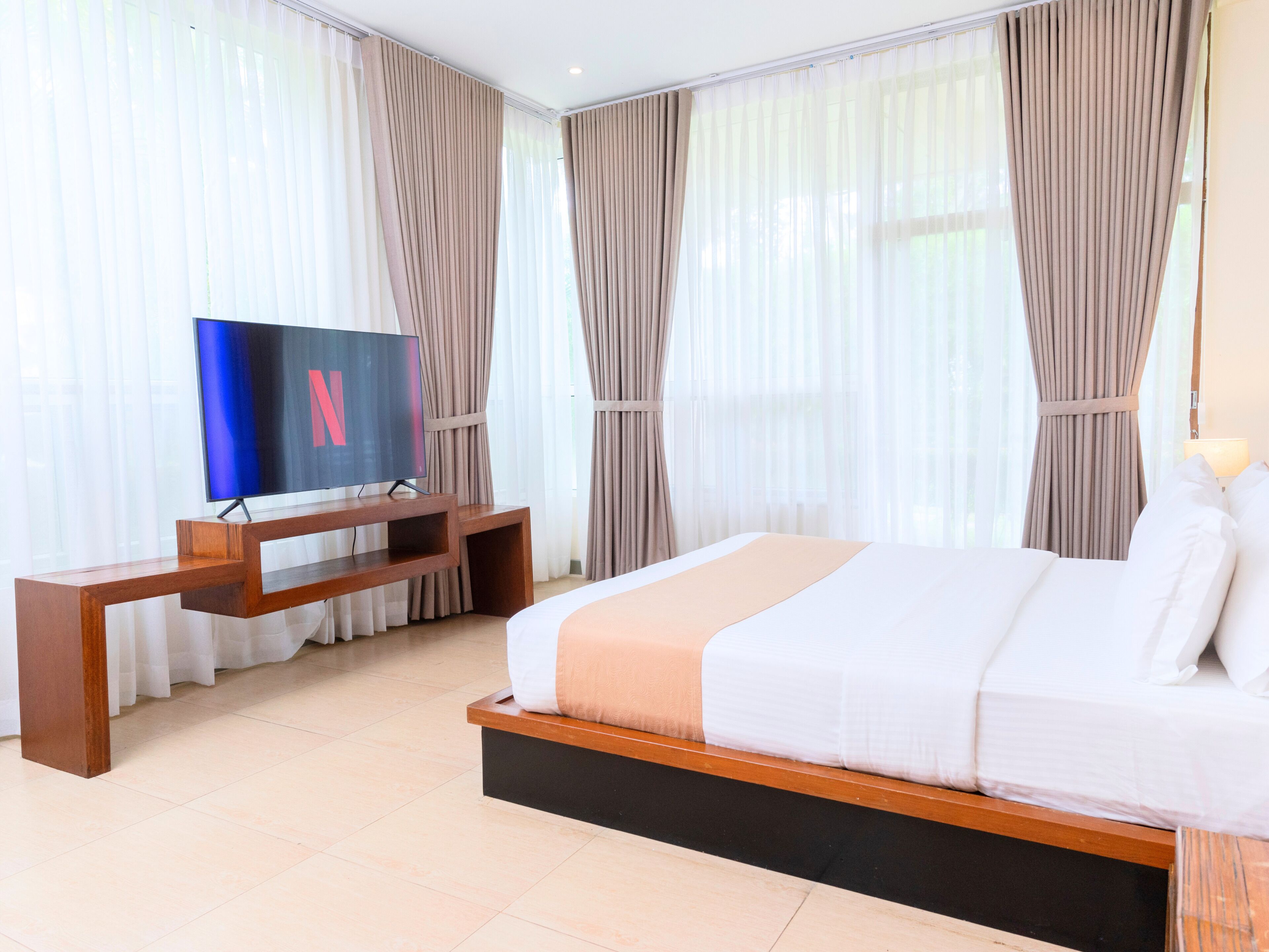 A hotel room with a bed, a TV on a wooden stand, and sheer curtains.