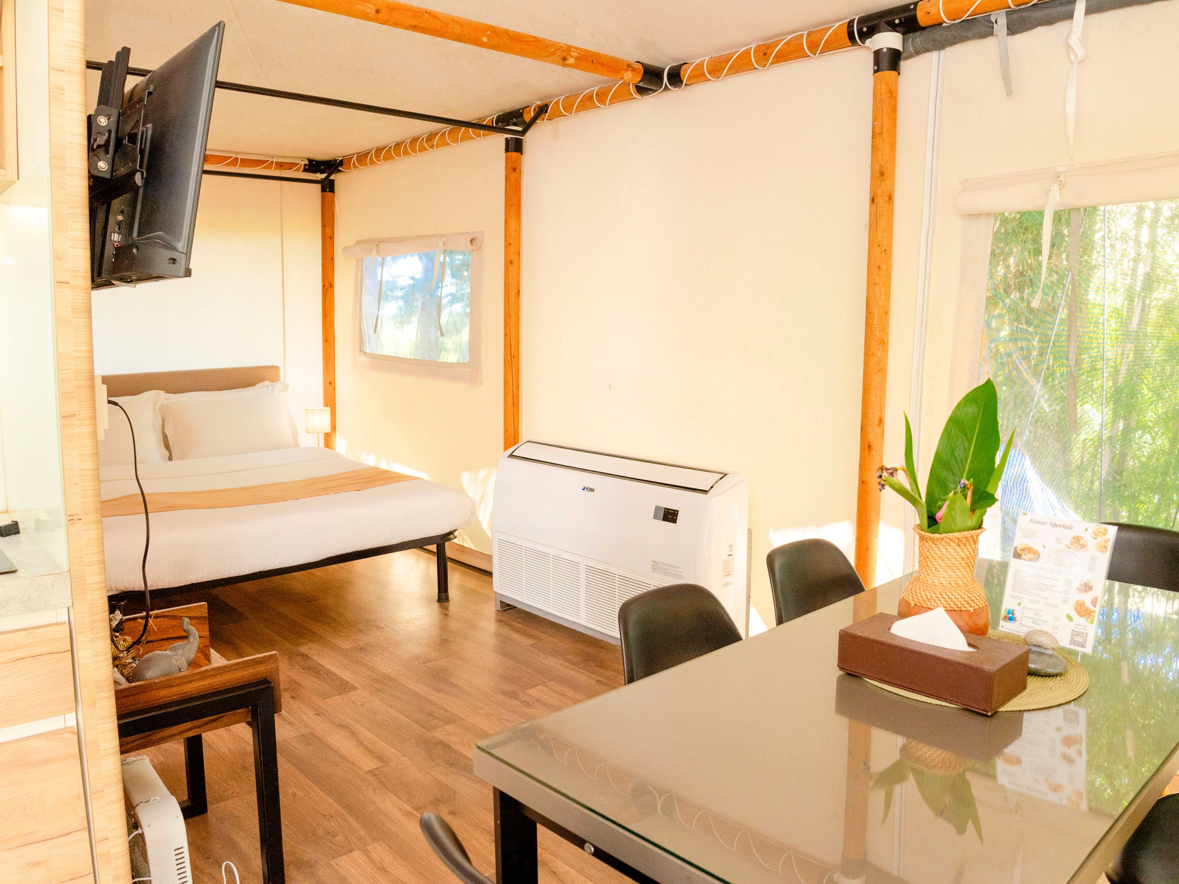 Habitación con cama, una mesa, sillas, un televisor montado en la pared, un aire acondicionado y una ventana con vistas a árboles.