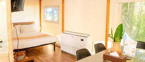 Habitación con cama, una mesa, sillas, un televisor montado en la pared, un aire acondicionado y una ventana con vistas a árboles.
